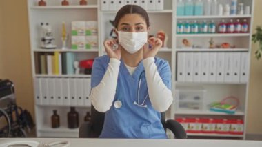 Mavi önlüklü İspanyol kadın doktor hastane kliniğinde maske takıyor ve kameraya gülümsüyor..