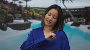 Mavi gömlekli kadın oteldeki havuz kenarında hareketler yapıyor. Etrafı tropikal bitkilerle çevrili ve rahatlatıcı bir ortam. Açık gökyüzünün altında canlı ve genç bir enerji yayıyor..