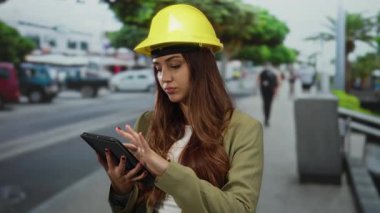 Sarı kask takan genç bir kadın dışarıda tablet üzerinde çalışıyor. Şehir merkezini ve mimari bağlamları gözler önüne seriyor. Mühendislik odaklı ve profesyonel çevreyi gösteriyor.