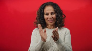 Neşeli kırmızı bir arka plana alkışlayan bir kadın, kıvırcık saçlar çerçeveli, rahat beyaz bir kazak giymiş, olgun güzelliğini ön plana çıkarıyor..