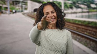 Kıvırcık saçlarıyla gülümseyen bir kadın, neşeli bir ifadeyle bir şehir caddesinde, demiryolu yakınında canlı ve canlı bir açık hava sahnesi yakalıyor..