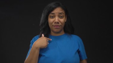 Stüdyoda siyah arka planda mavi gömlek giyen Afro-Amerikalı kadın parmak göğse işaret ediyor. Kendinden şüphe ediyor..