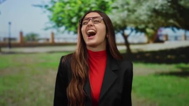 Gözlüklü bir kadın parkta gülümsüyor ve bağırıyor, kırmızı bir bluz ve siyah bir ceket giyiyor, ağaçlarla çevrili güneşli bir günde neşe saçıyor..