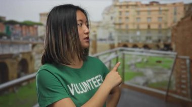 Bir şehir caddesindeki Roma harabelerini işaret eden gönüllü bir kadın, açık hava ortamında yeşil bir tişört giyiyor, gençliği ve Asya kökenliyi temsil ediyor..