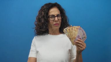 Orta yaşlı İspanyol kadın Meksika Pezosu 'nu elinde tutuyor ve mavi stüdyoda kameraya yumruğunu uzatıyor..