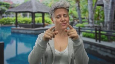 Gümüş saçlı kadın, havuz alanındaki izleyiciye iki eliyle işaret ediyor..