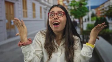 Gün ışığıyla aydınlatılmış ağaçlarla ve kentsel binalarla dolu bir sokakta şaşırdığını ifade eden gözlüklü İspanyol bir kadın.