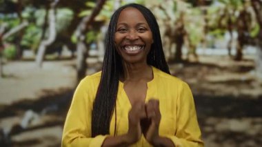 Parkta ışıl ışıl gülümseyen bir kadın. Arka planda ağaçlar olan sarı bir gömlek giyiyor. Güneşli bir günde neşe ve doğa sergiliyor..