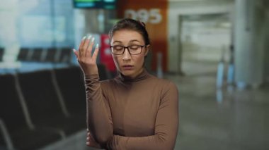 Havaalanı terminalinde gözlüklü bir kadın düşünceli görünüyor, ayrılış bölgesindeki bir yansımayı vurguluyor..