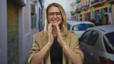 Gözlüklü gülümseyen sarışın kadın park halindeki arabalarla ve renkli binalarla dolu kalabalık bir şehir caddesinde rahatça duruyor..