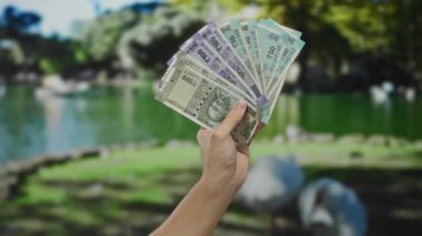Parkta elinde Hint banknotları tutan beyaz bir adam, arka planda yemyeşil bir gölet ve yemyeşil bir ortamda mali başarı gösteriyor..