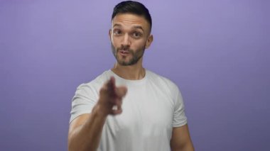 Latin asıllı genç bir adam, mor bir arka plana karşı ciddi bir ifadeyle itaatkar bir jest yapıyor, beyaz gömlek giyiyor ve güçlü bir mesaj ima ediyor..