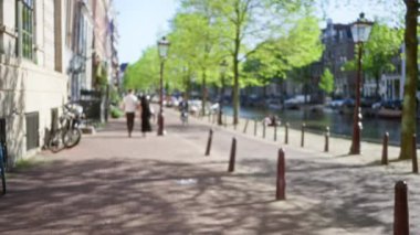 Odaklanamayan bir sahne, canlı, kentsel bir manzaranın özünü somutlaştıran yemyeşil ağaçlar, bisikletler ve tarihi binalarla Amsterdam Kanalları 'nın yanında yürüyen bir erkek ve kadını yakalar..