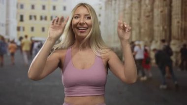 Sarışın, gülümseyen kadın kollarını kaldırıp dans ediyor ve neşeli bir yüz ifadesiyle sokakta kutlama yapıyor..