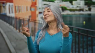 Gri saçlı kadın deniz kenarındaki gezinti alanında anlamlı jestler yapıyor, mavi kazak giyiyor, sahil manzarası ile çevrili, açık hava özgürlüğü ve hareketli ortamda meydan okumayı temsil ediyor..