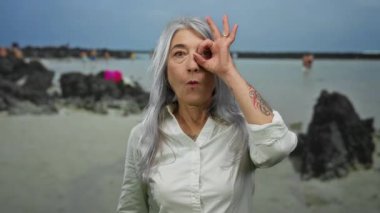 Üst düzey bir kadın, kır saçlı ve neşeli bir ifadeyle manzaralı bir sahil sahilinde dikilip açık havanın tadını çıkarırken, gözünün önünde iyi bir jest yapıyor.