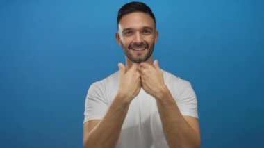 Beyaz tişörtlü genç İspanyol adam ellerini kameraya doğru uzatıyor, mavi bir arka plana gülümsüyor, dostça ve samimi bir jest yapıyor..