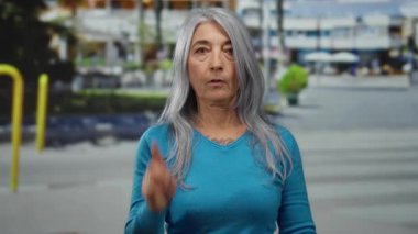 Son sınıf öğrencisi kadın sokakta jest yapıyor, mavi bir süveter giyiyor kararlı bir ifadeyle, çevresi binalar ve yeşilliklerle çevrili..