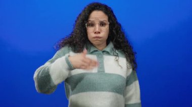 Gözlüklü ve çizgili süveter giyen genç İspanyol kadın, izole edilmiş mavi arka plan duvarından endişe ederek ağzını kapatıyor..