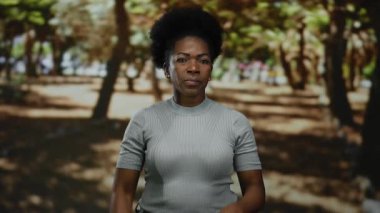 Parkta ağaçlarla yemin eden, ciddi bir ifade sergileyen, açık havada giyinen bir kadın..