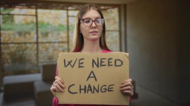 Elinde karton tabela olan genç sarışın kadın stüdyoda bir değişikliğe ihtiyacımız var; sosyal aktivizm dayanışması umudu.