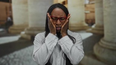 Gözlüklü kadın şehir sokağında kendini hasta hissediyor. Kırmızı gözlüklü, çizgili tişörtlü, kıvırcık saçlı, kenar mahalleli bir kadın..