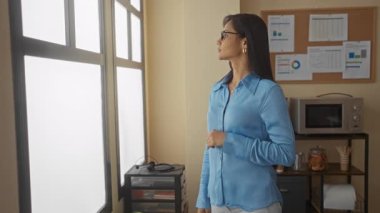 Gözlüklü ve mavi tişörtlü genç bir kadın, doğal ışık ve ergonomik mobilyalarla modern bir çalışma ortamını ön plana çıkarıyor..