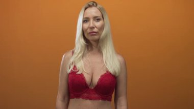 Stüdyoda duran sarışın kadın turuncu bir arka planda kırmızı dantel sutyen dekoltesi gösteriyor..