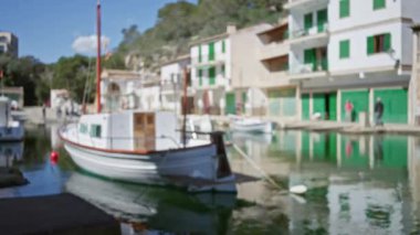 Mallorca 'daki Cala figera limanının bulanık görüntüsü, dağılmış tekneler ve insanlar, geleneksel Akdeniz mimarisinde sıcak güneş ışığı canlı renkleri ve sakin atmosferi vurguluyor.