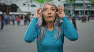 Gri saçlı bir kadın, kalabalık bir şehir caddesinde, açık havada, insanlarla çevrili, şehir ortamında duygu ve titreşim sergiliyor..