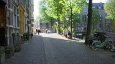 Bisikletli bulanık şehir manzarası ve güneşli bir günde gölgelerini düşüren tarihi binalar ve yemyeşil ağaçlarla dolu bir Avrupa caddesi boyunca yürüyen insanlar..