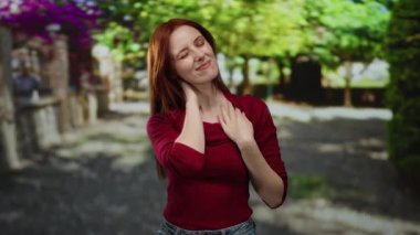 Güneşli bir şehir caddesinde dışarıda duran bir kadın kızıl saçlı ve günlük kıyafetleriyle boyun ağrısı çekiyor. Arka planda bulanık yeşillik ve canlı çiçeklerle çevrili.