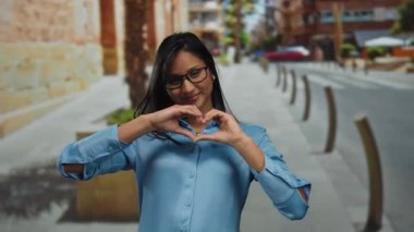 Dışarıda gülümseyen bir kadın, güneşli caddede elleri gözlüklü ve mavi gömlekli kalp yapıyor.