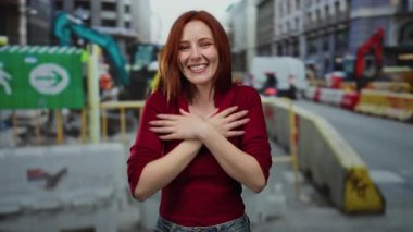 Şehir merkezinde kızıl saçlı, kırmızı kazaklı bir kadın inşaat alanında gülümsüyor canlı enerji ve şehir manzaralı modern gelişimi sergiliyor..