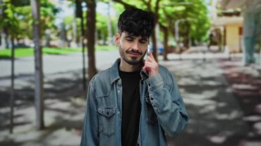 Kot ceketli genç adam şehir sokağında gün boyunca yapraklı ağaçların altında sessizlik için el kol hareketi yapıyor. Şık ve açık hava manzarasında sakin ve yakışıklı özellikler sergiliyor..