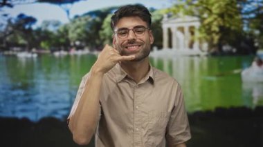 Latin kökenli bej gömlekli genç adam yeşil parkta telefonla jest yapıyor. İletişim daveti dostane..