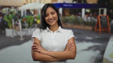 Sokak restoranı ve yeşilliğin bulanık arka planına sahip açık bir kafede kollarını kavuşturarak güvenle gülümseyen genç bir kadın..