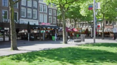 Amsterdam şehir sokak sahnesi bulanık kafeler, ağaçlar ve binalar arka planda bir bokeh etkisi yaratıyor, canlı şehir atmosferini vurguluyor.