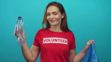 Genç gönüllü kadın plastik şişe ve mavi geri dönüşüm çantasını turkuaz stüdyo duvarına kaldırıyor; sürdürülebilirlik aktivizmi.