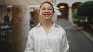 Yüzü gülen beyaz gömlek giyen ve restoran terasında taş sütunlarla çerçevelenmiş gözleri kapalı bir kadın; huzur yansıması.