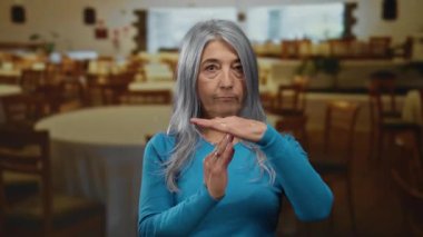 Son sınıf öğrencisi, gri saçlı, kapalı bir restoranda mola jesti yapıyor. Arka planda ahşap sandalyeler ve masalar olan mavi bir süveter giyiyor..