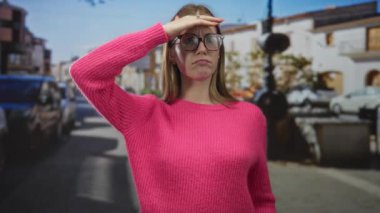 Kadın yuvarlak gözlük arkasında gözleri kalkan ve güneşli bir sokakta açık avuç işaret; davet ve merak.