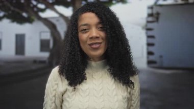 Beyaz süveter giyen Afro-Amerikalı bir kadın kamerayı binanın önünde işaret ediyor..