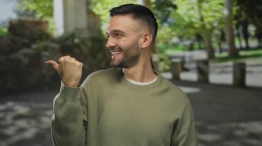 Yeşil parkta gülümseyen İspanyol bir adam. Parmakla dışarıyı işaret ediyor. Güneş ışığı, yemyeşil ağaçların arasından süzülüyor..