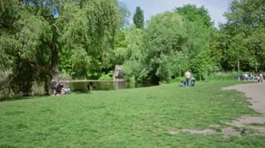 Bulanık park sahnesi, insanların ağaçların altında güneşli bir günde ağaçların altında rahatlayıp yeşilliğin tadını çıkardıkları, boş zamanların ve doğanın altını çizdiği güneşli bir gün..