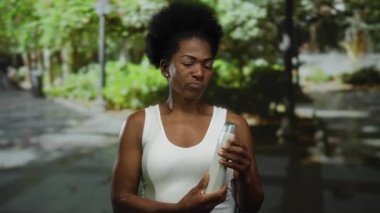 Parkta şampuan tutan bir kadın, günlük giysiler giymiş, belirsizliğini dile getirirken yemyeşil bir ortam içinde..