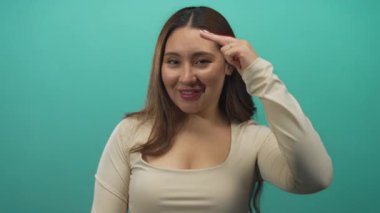 Latin kökenli genç bir kadın turkuaz stüdyoda kameraya çıplak elle gülümser ve işaret eder..