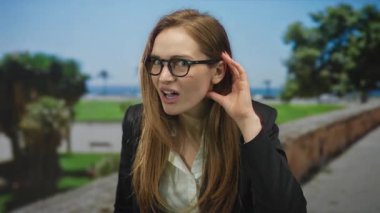 Siyah gözlüklü bir kadın kulağını güneşli bir sokakta palmiye ağaçları ve tuğla duvarın yanındaki kaldırıma dayadı..