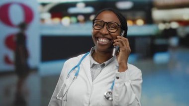 Beyaz önlüklü, steteskopla gülümseyen bir kadın kapalı hava alanında akıllı telefondan konuşuyor. Profesyonel iletişim ve seyahat temasını öneriyor..