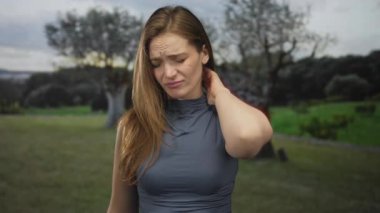 Kolsuz gri bluzlu kadın elini zeytin ağaçlarıyla dolu yeşil bir parkta boynuna bastırıyor..